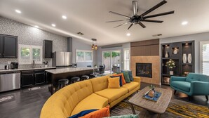 A modern kitchen with stainless steel appliances, a dining area with a curved yellow sofa, and a living room with a fireplace and teal armchairs. - Welcome to Villa Tempo - Pool, Spa, Game Room, BBQ, Fast Wifi, Sleeps 12+ (Coachella)
