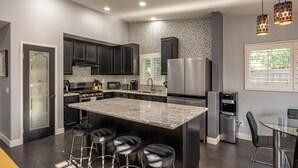 A modern kitchen with dark cabinets, a granite island, stainless steel appliances, and a bar area with stools. - Welcome to Villa Tempo - Pool, Spa, Game Room, BBQ, Fast Wifi, Sleeps 12+ (Coachella)