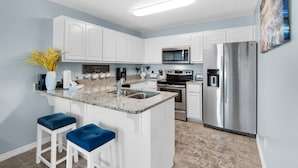 A modern kitchen with white cabinetry, a granite countertop, stainless steel appliances, and blue bar stools. - Gulf Island 232 - Sunset Suite (Navarre)