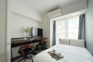 A hotel room with a bed, a desk with a chair, a TV, and an air conditioning unit.