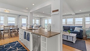 A modern kitchen with a central island, a dining area with chairs, and a living room with a sofa and a view of the sea.