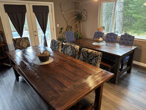 Uma mesa de jantar de madeira com cadeiras estampadas, uma cesta trançada e uma janela com vista para árvores.