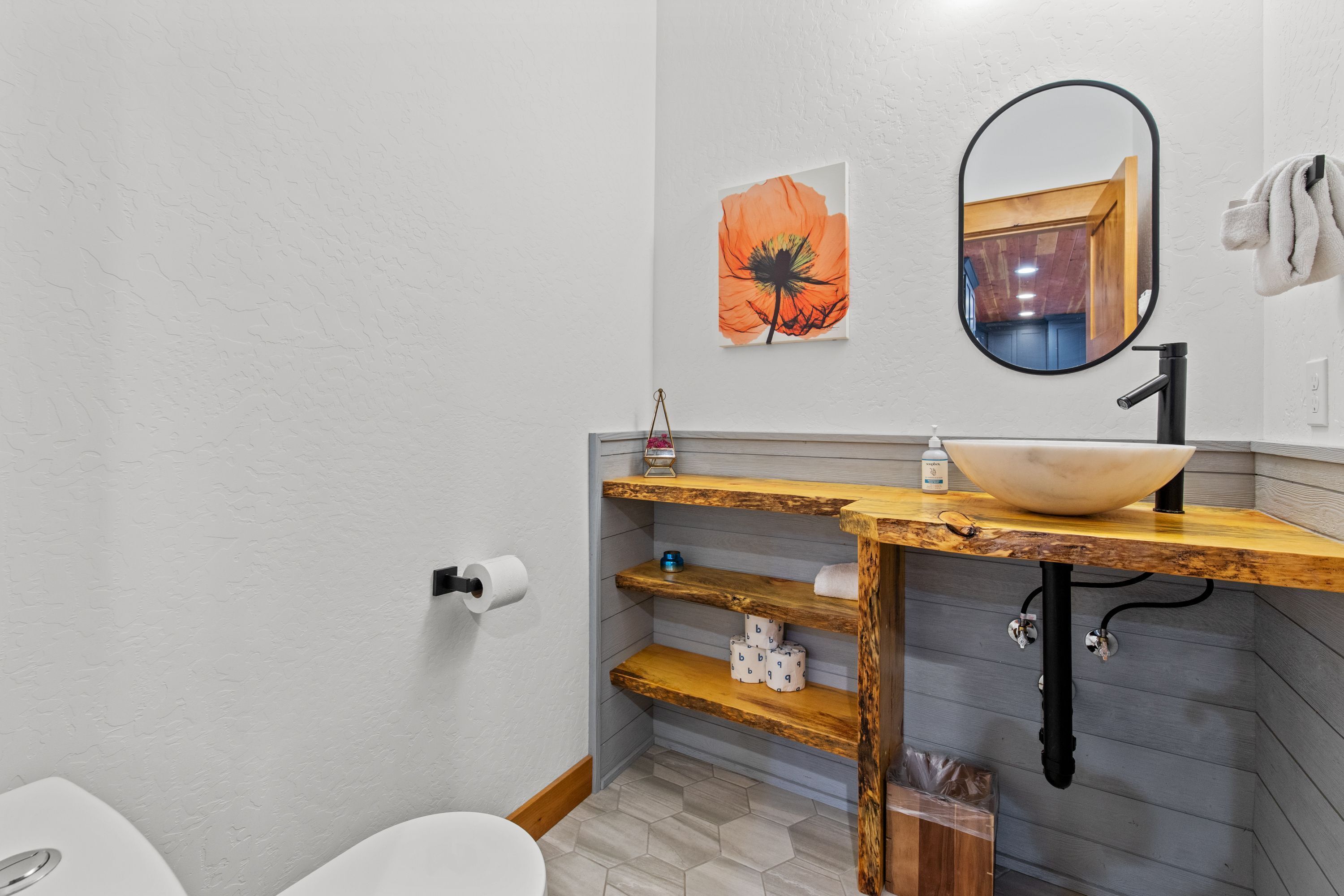 Une salle de bain avec des toilettes blanches, un meuble-lavabo en bois doté d’un grand miroir et un lavabo fixé au mur.