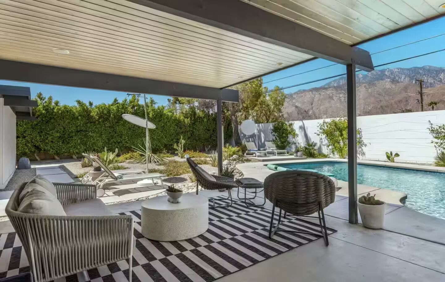 Une terrasse extérieure avec une piscine, aménagée avec des fauteuils en osier et une table ronde, le tout entouré d’un tapis à carreaux.
