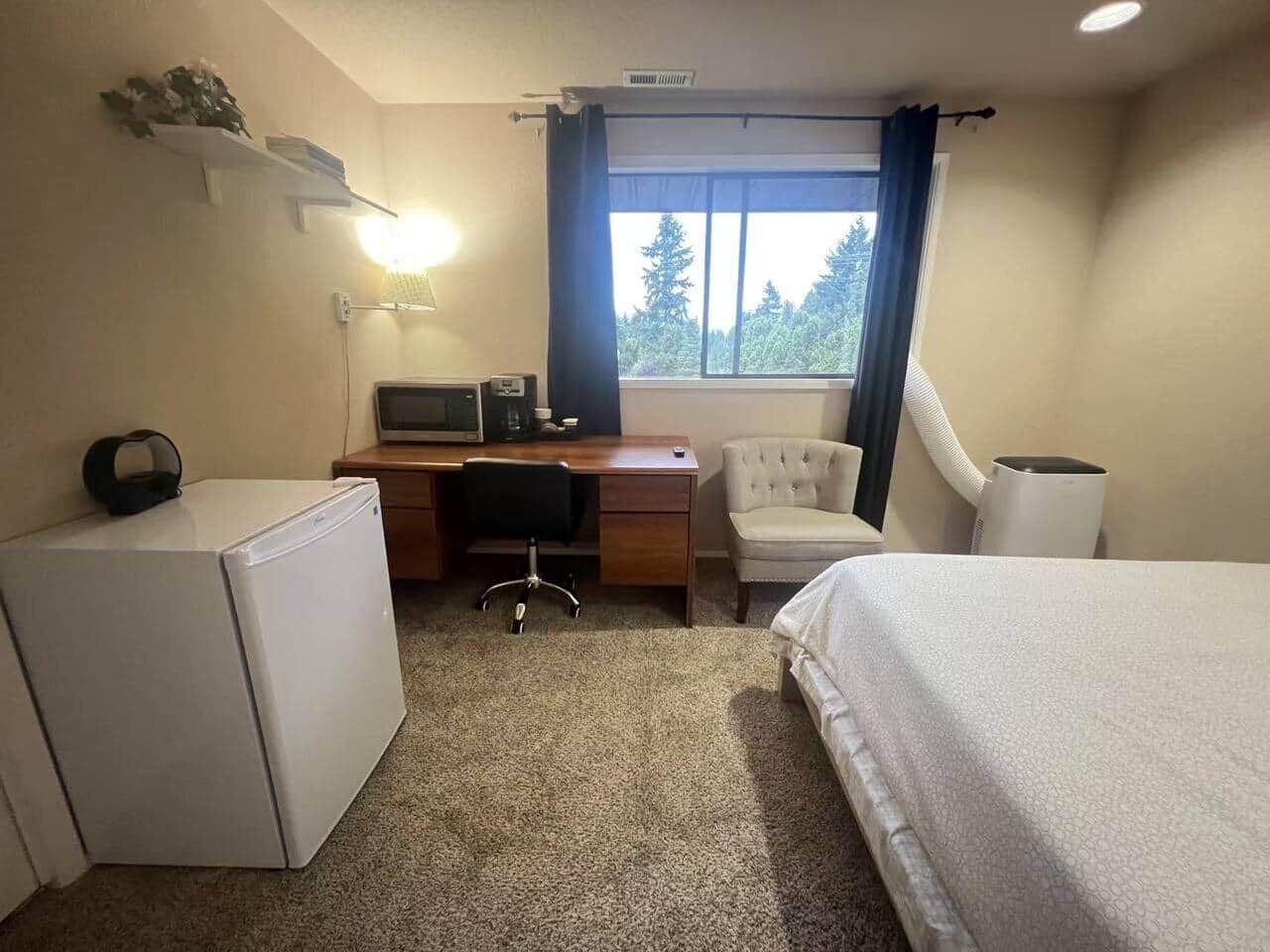 A hotel room with a bed, a desk with a computer, a chair, a small refrigerator, and a window with curtains.