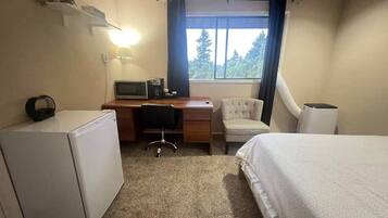 A hotel room with a bed, a desk with a computer, a chair, a small refrigerator, and a window with curtains.