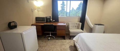 A hotel room with a bed, a desk with a computer, a chair, a small refrigerator, and a window with curtains.