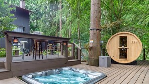 A hot tub on a wooden deck surrounded by trees, a wooden sauna, and a covered seating area with chairs and a table. - Crystal Dome · Private Glamping Dome w/ Hot Tub, Sauna & Panoramic View (Eastmont)