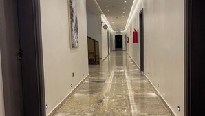 Un pasillo largo y bien iluminado con suelo de mármol, puertas a ambos lados y un extintor en la pared derecha.