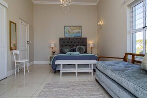 Quarto com cama, um banquinho, uma cadeira, uma janela e um quadro na parede.