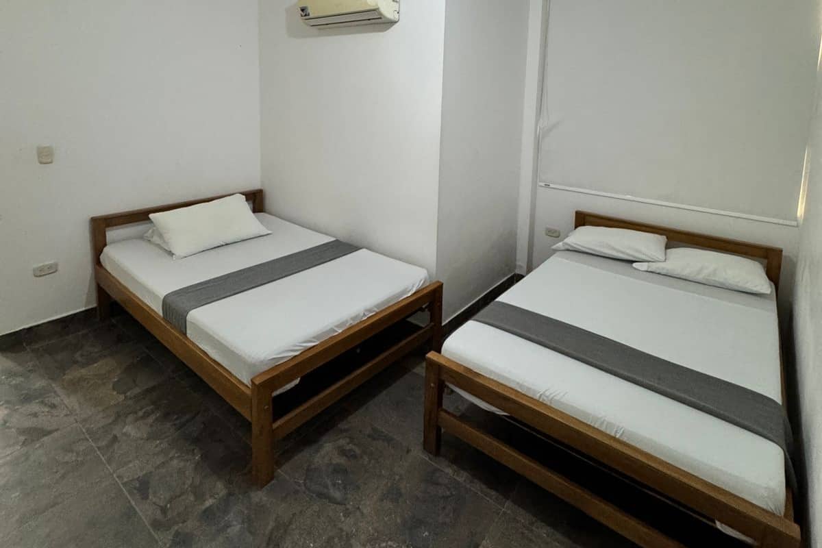 A small, clean room with two wooden beds, white walls, a ceiling light, and a wall-mounted air conditioner.