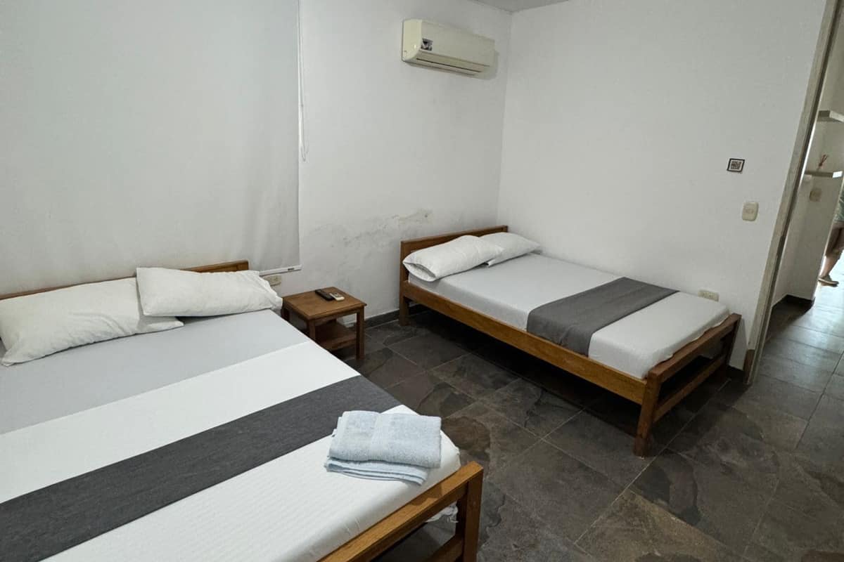 A room with two beds, a bedside table, an air conditioning unit, and a tiled floor.