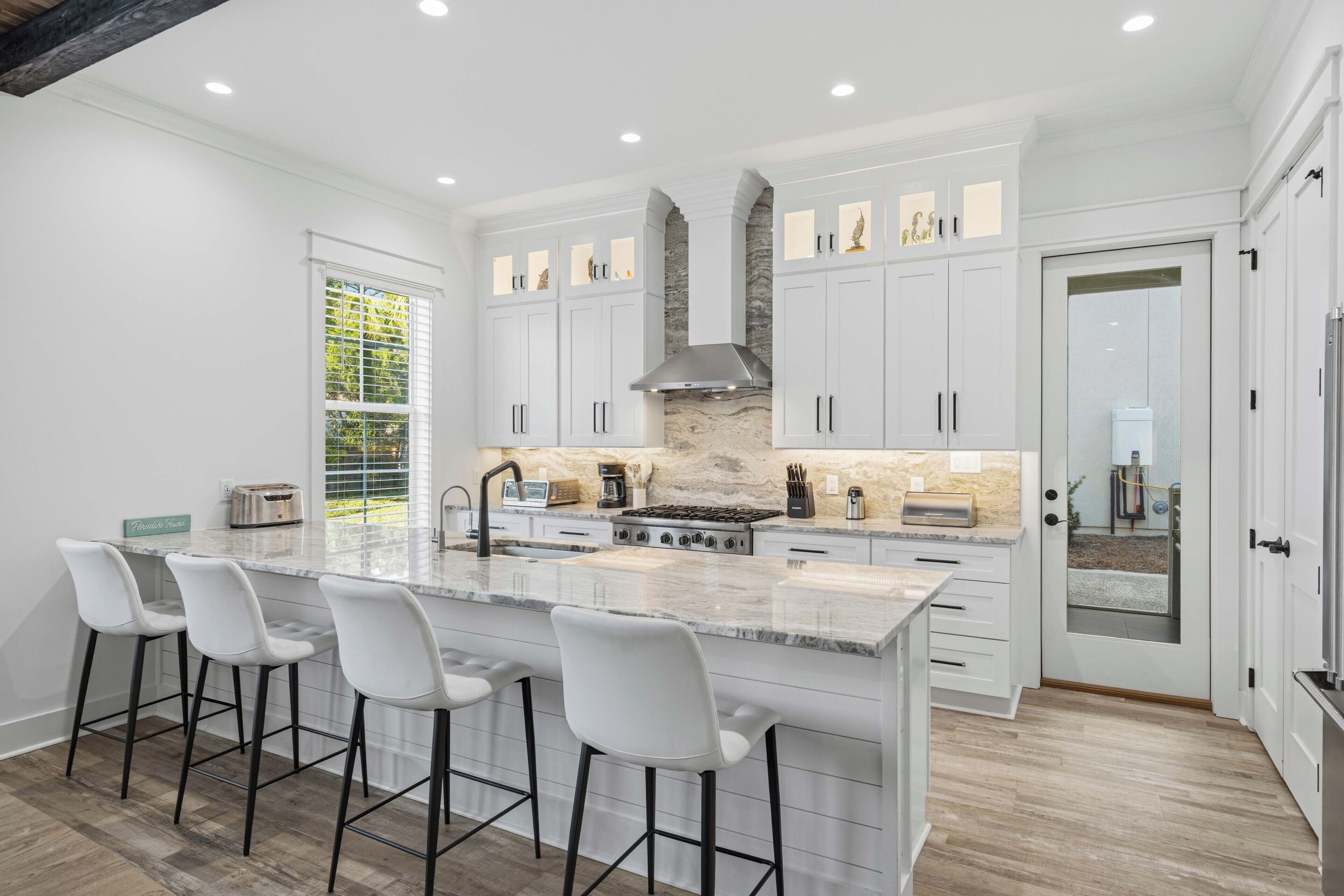 A modern kitchen with white cabinetry, a large island with bar stools, a stone backsplash, and recessed lighting.