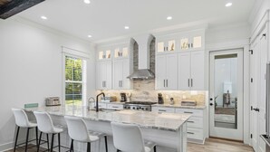 Una cocina moderna con armarios blancos, una isla grande con taburetes de bar, salpicadero de piedra e iluminación empotrada.