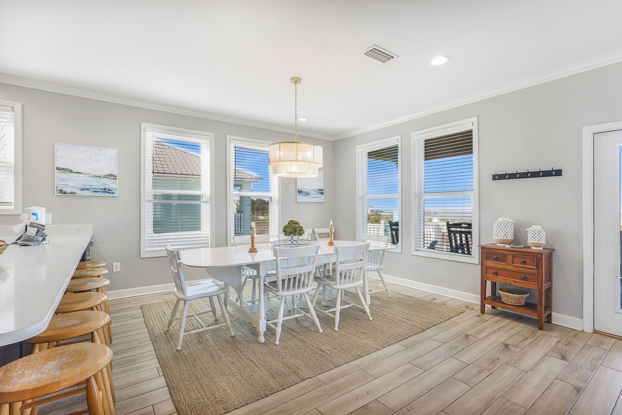 A modern kitchen with a white island, bar stools, a dining table with chairs, and large windows with blinds.