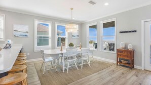 A modern kitchen with a white island, bar stools, a dining table with chairs, and large windows with blinds. - 1 BLOCK to the BEACH! Weekly/Monthly promotions! Bay views! Dog friendly! (Gulf Shores)