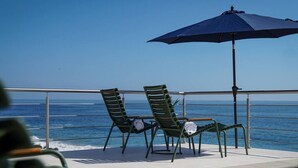 Eine Terrasse mit zwei Stühlen, einem Tisch und einem Sonnenschirm, mit Blick auf den Ozean.