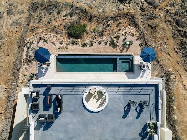 Vista aérea de un área de piscina en la azotea con sillones de descanso y sombrillas, rodeada por un paisaje rocoso.