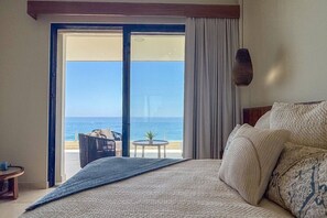 Une chambre d’hôtel avec un lit, une vue sur la mer, un balcon avec une table et des chaises, et une fenêtre avec des rideaux.