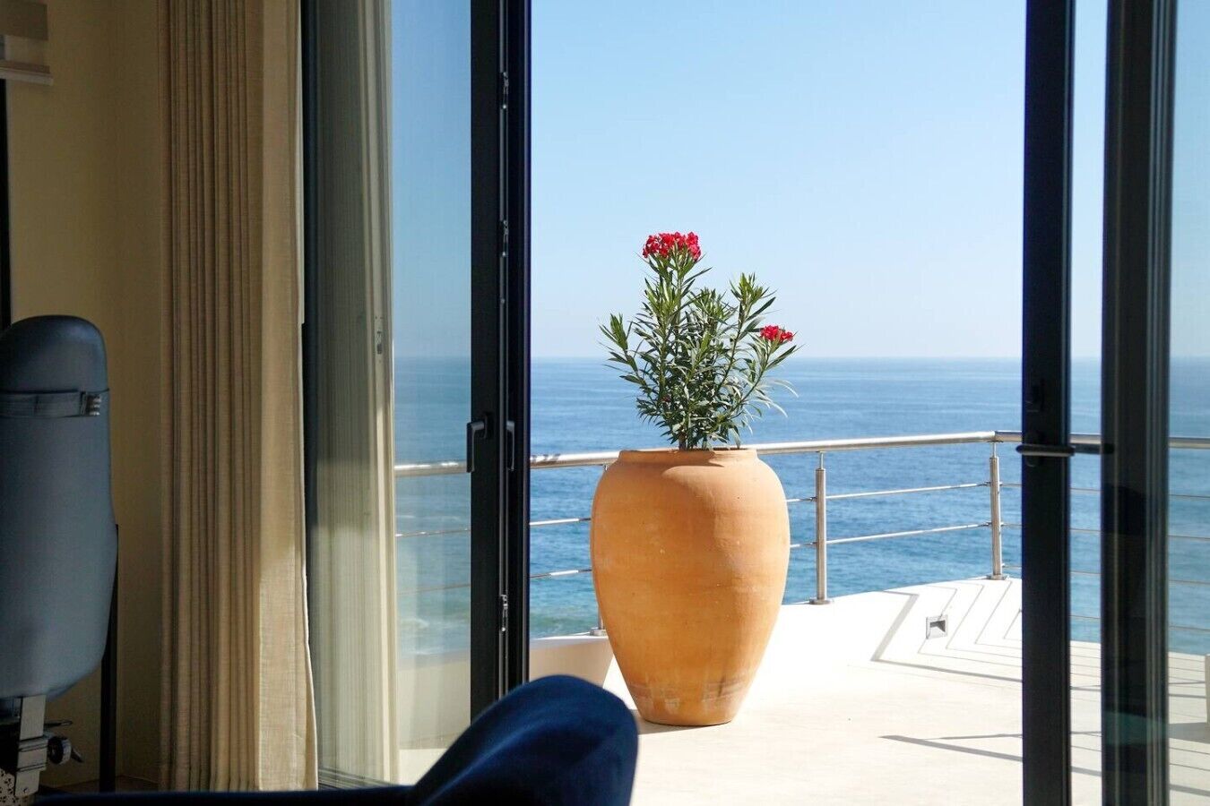 A balcony with a large terracotta pot containing red flowers, overlooking the sea.