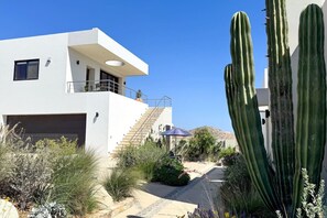 Ein modernes, zweistöckiges Gebäude mit flachem Dach, einer Treppe, die zum oberen Stockwerk führt, und einem Gartenbereich mit Kakteen und Wüstenpflanzen.