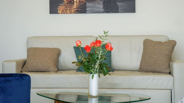 Un salon avec un canapé beige, deux coussins, une table basse en verre et un vase rempli de fleurs rouges.