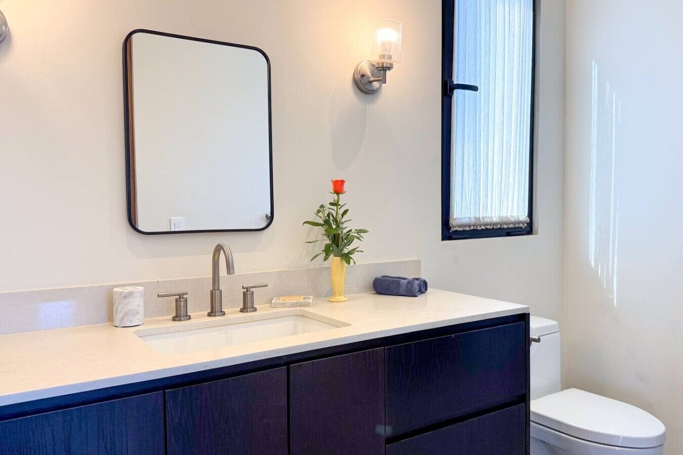 A bathroom with a white sink, a mirror, a toilet, and a window with blinds.