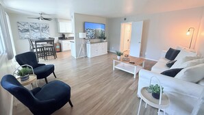 Uma sala de estar moderna com um sofá branco, uma mesa de centro e uma poltrona azul. Há uma área de jantar com mesa e cadeiras, e uma cozinha ao fundo.