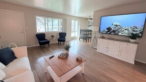 Uma sala de estar espaçosa com uma mesa de centro de madeira, duas poltronas, um sofá, uma área de refeições com mesa e cadeiras, e uma grande TV de tela plana sobre um móvel branco.