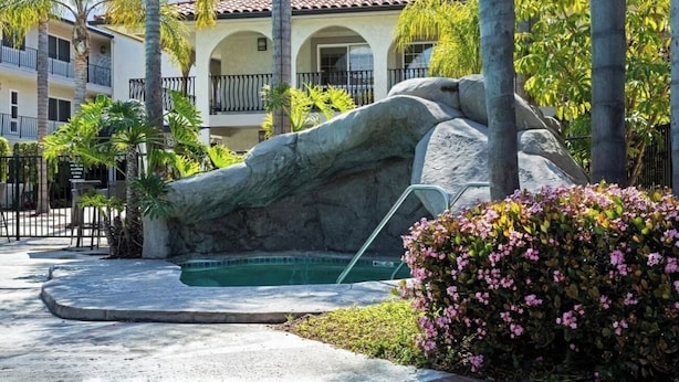 Área de piscina com uma formação rochosa, palmeiras e um edifício com telhado telhado.