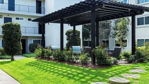 Uma área de estar coberta ao ar livre, com pérgola, rodeada por relva e arbustos bem cuidados, e adjacente a um edifício com varandas.