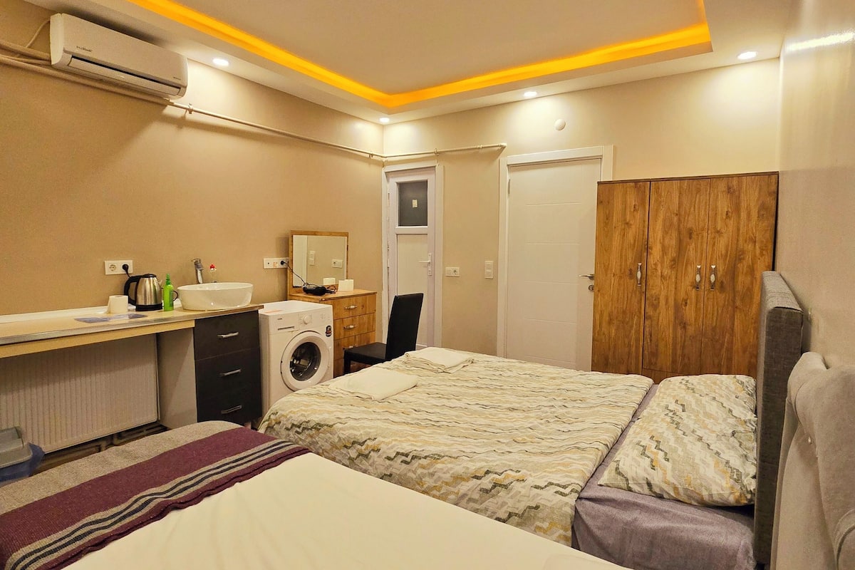 A hotel room with a bed, a washing machine, a small desk with a kettle, and a wardrobe.