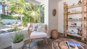 A cozy reading nook with a wicker chair, a small round table, and a bookshelf filled with books and decorative items. - Palm Springs Oasis • 3 King Ensuites + Pool & Spa (Palm Springs)