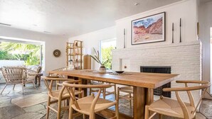 A dining area with a wooden table and woven chairs, a white brick fireplace, and a bookshelf. - Palm Springs Oasis • 3 King Ensuites + Pool & Spa (Palm Springs)