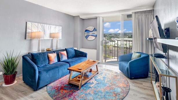 A modern living room with a blue sofa, a wooden coffee table, and a colorful rug. There is a balcony with a view of the city. - Hilo Lagoon Center (Hilo)