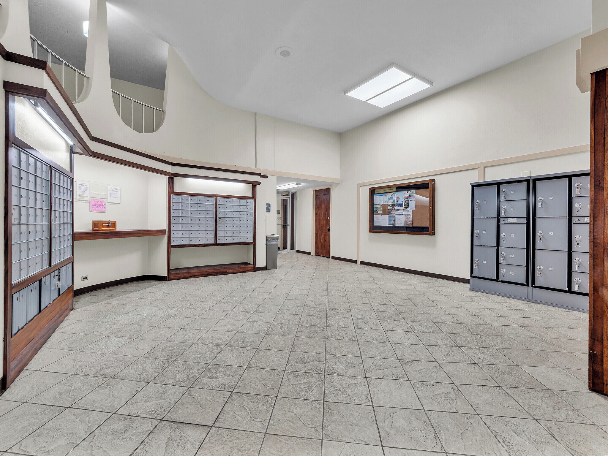 A spacious hallway with a tiled floor, a wooden bench, a bulletin board, and a row of metal lockers.
