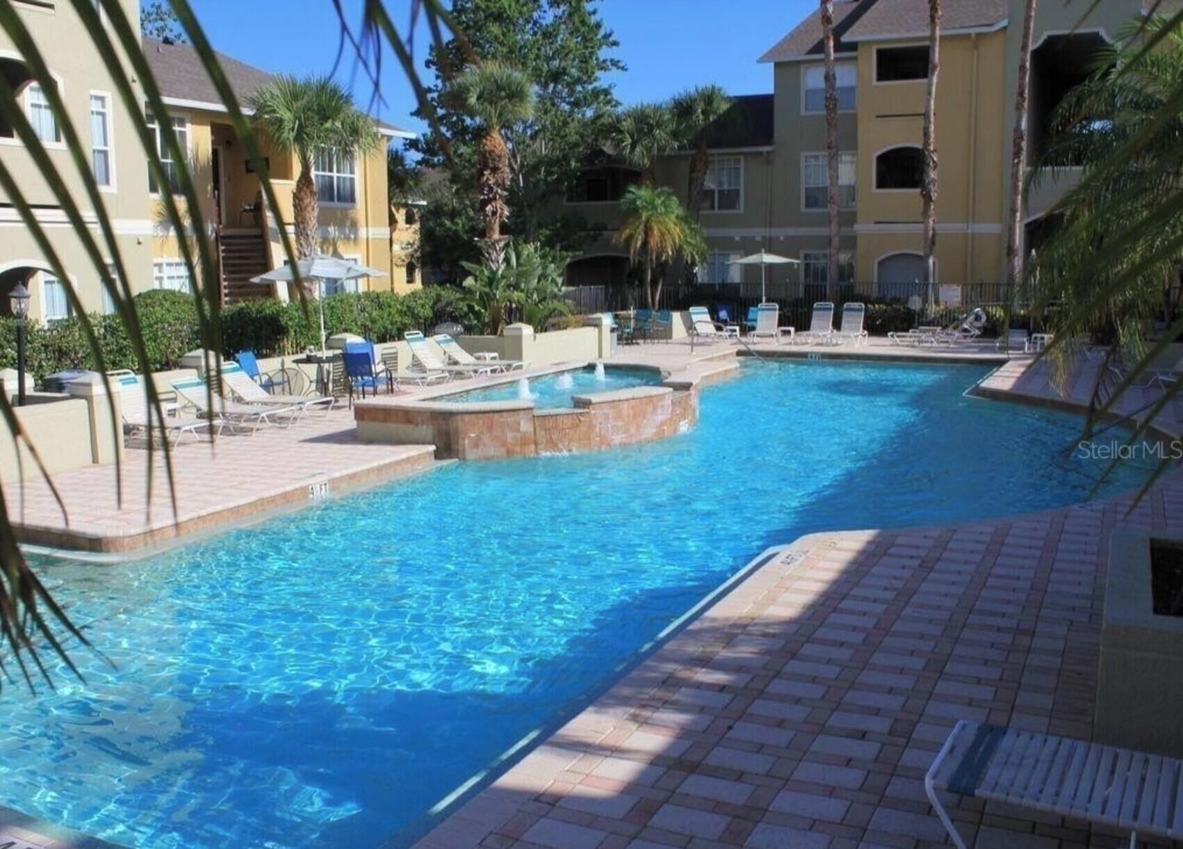 A swimming pool with lounge chairs and a small hot tub, surrounded by buildings with balconies and palm trees.