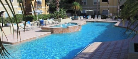 A swimming pool with lounge chairs and a small hot tub, surrounded by buildings with balconies and palm trees.