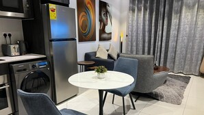 A modern kitchen with a built-in oven, a refrigerator, and a washing machine. There is a round dining table with two chairs and a small potted plant. - Chic 1-bedroom apartment at The Lennox, Airport Residential Area (Accra)