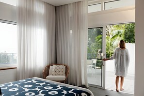 Une chambre d’hôtel avec un lit, un fauteuil en osier, un balcon donnant sur un paysage verdoyant, et une personne qui ouvre une porte coulissante.