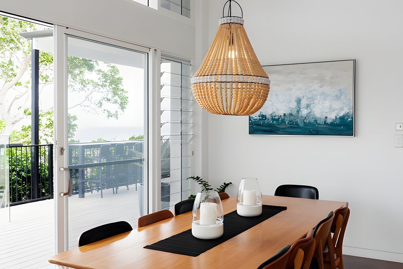 Un coin repas avec une table en bois, des chaises, une suspension en fibres tressées, un vase blanc et un tableau représentant un paysage urbain accroché au mur.