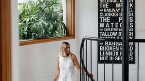 Una mujer con vestido blanco bajando una escalera con barandas negras, una ventana con follaje visible, y una pared con texto negro.
