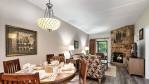 A dining area with a round table set for six, flanked by wooden chairs. A stone fireplace with a television is visible in the background.