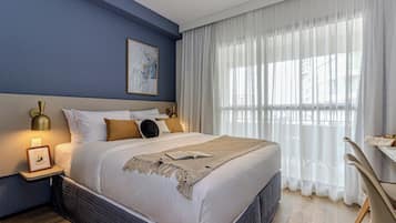 Un dormitorio ordenado con una cama grande, una mesita de noche con una lámpara, un cuadro enmarcado y una ventana con cortinas translúcidas.