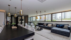Ein modernes Wohnzimmer mit einem großen dunklen Holztisch, weißen Stühlen, einem grauen Sofa mit blauen und beigen Kissen sowie hängenden Pendelleuchten.