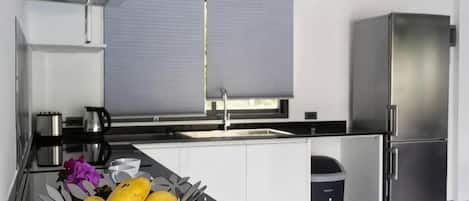 A modern kitchen with a stainless steel refrigerator, a sink, and a window with blinds.