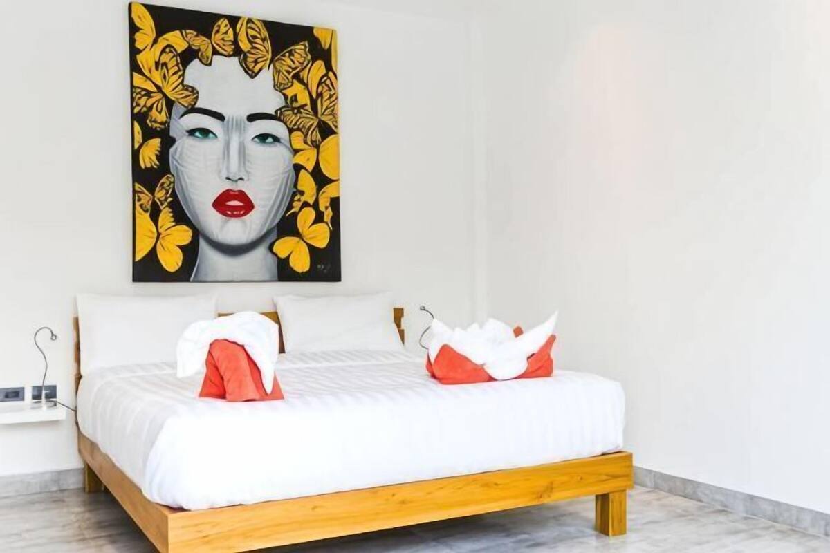 A bed with white bedding and red pillows, a wooden headboard, and a painting above the bed.