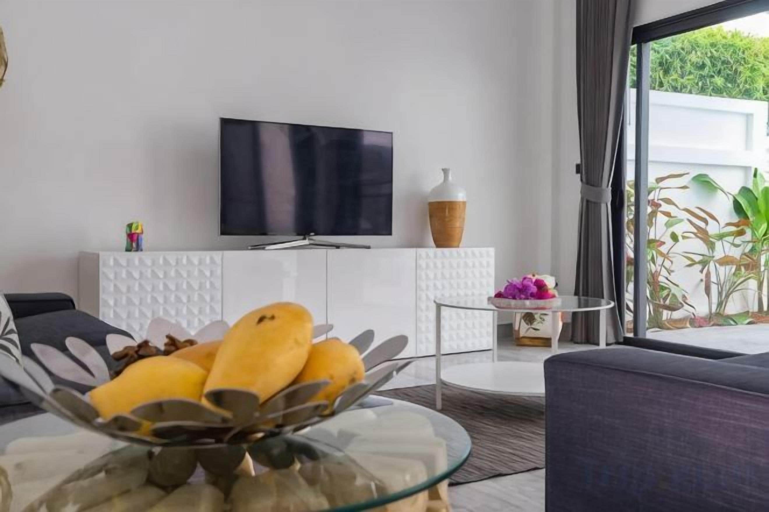 A modern living room with a flat-screen TV, a glass coffee table with fruit, and a view of greenery outside.
