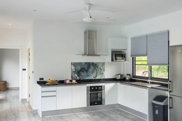 A modern kitchen with white cabinets, a black countertop, a stainless steel range hood, and a window with blinds. - Villa Mokkaran, 3 Bedrooms (Koh Samui)