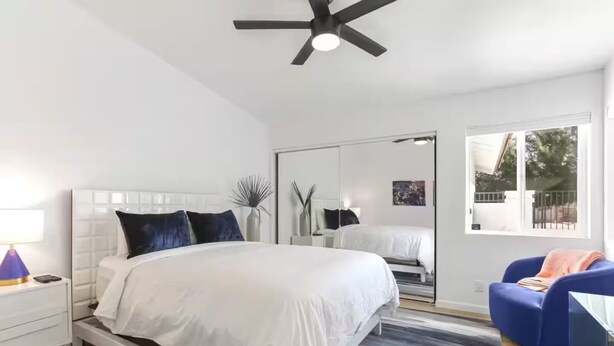 A modern bedroom with a large bed, a ceiling fan, a nightstand, a mirror, and a window with a view of the outside. - Palm Springs Pool Home - Spa + Family Retreat (Palm Springs)